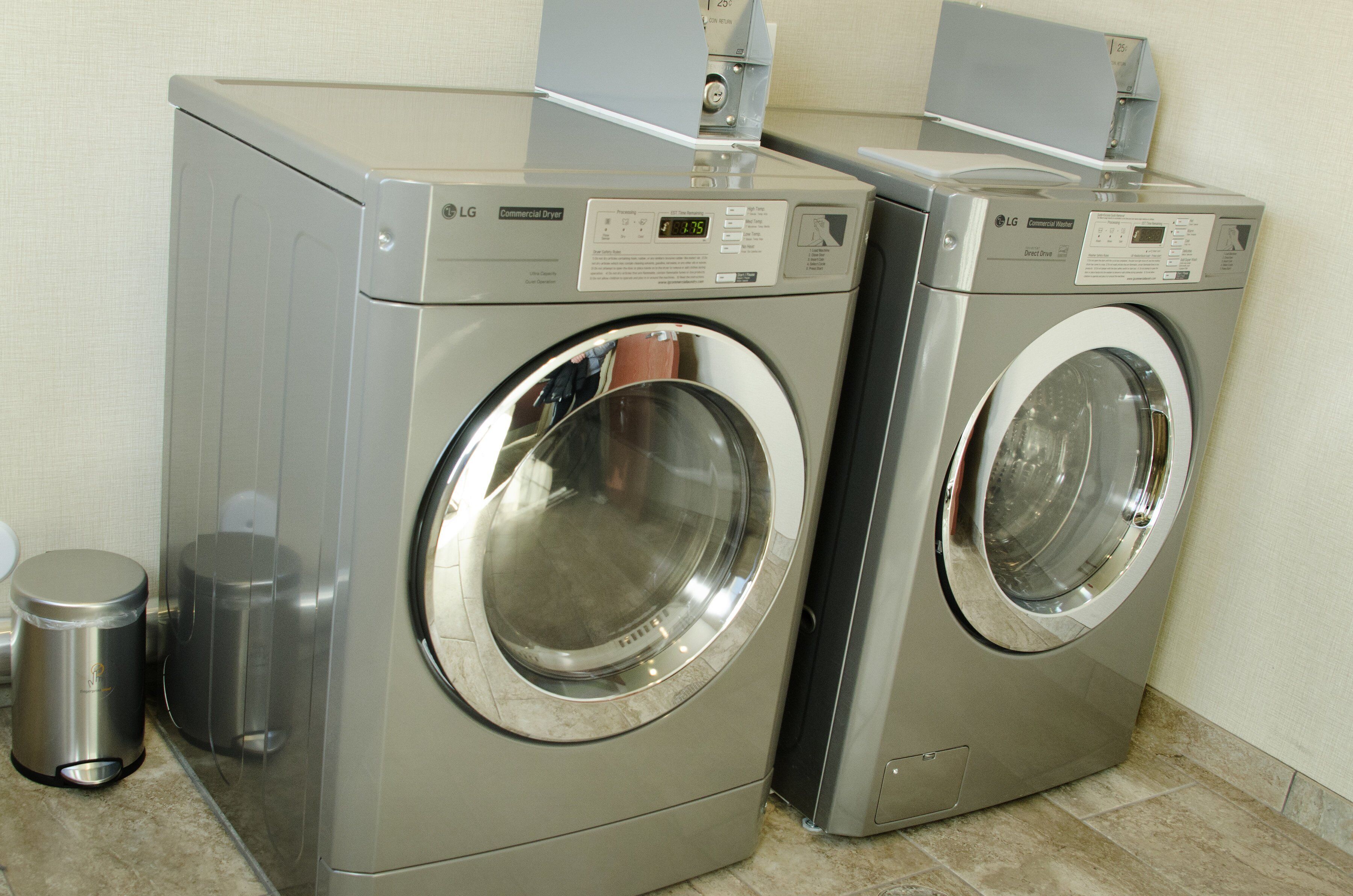 Two commercial dryers in a laundry room with a trash can and a tiled floor.