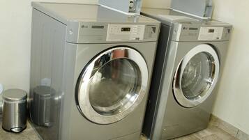Two commercial dryers in a laundry room with a trash can and a tiled floor.