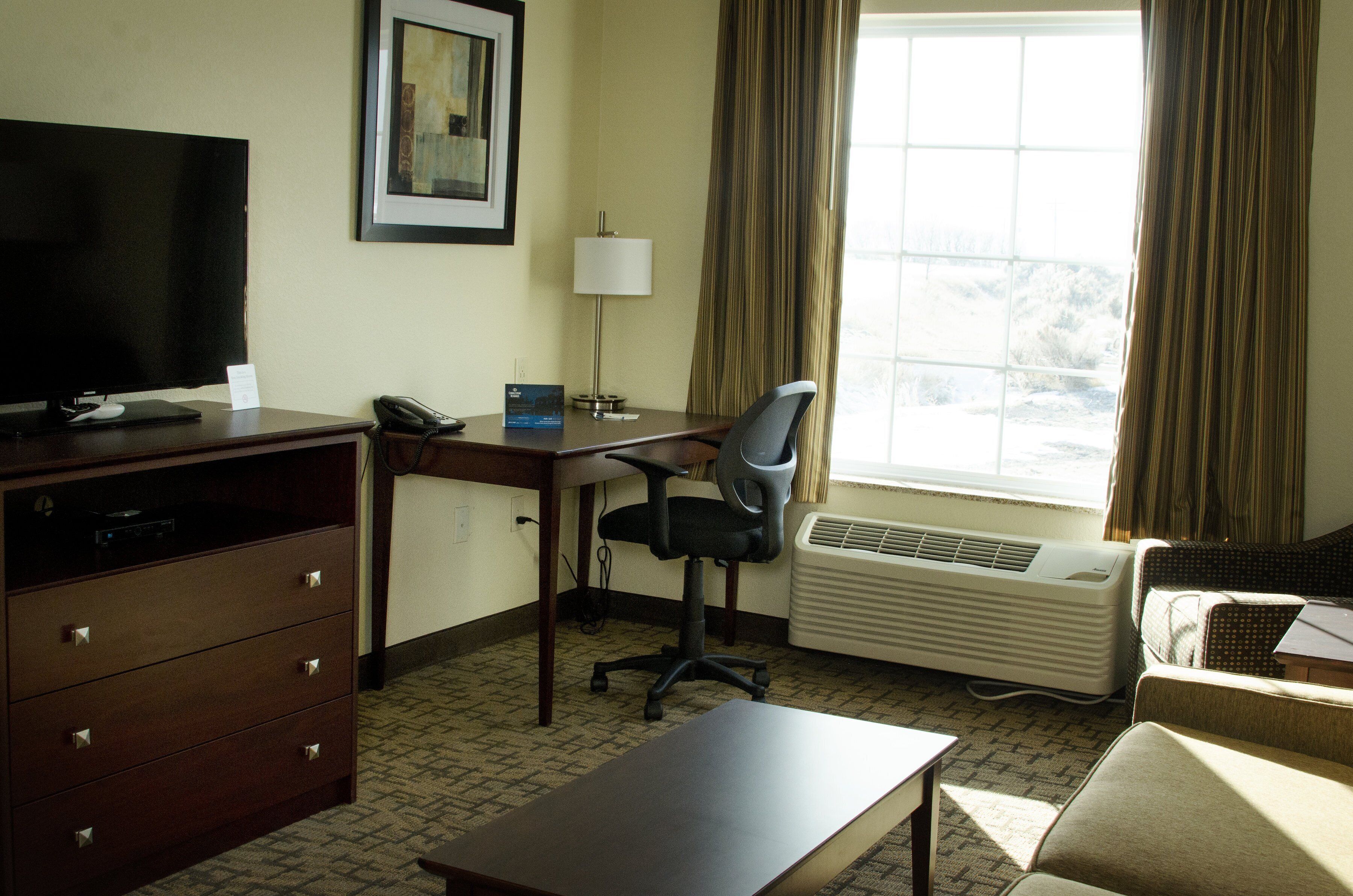 A hotel room with a desk, chair, television, air conditioner, and a view of the outdoors.