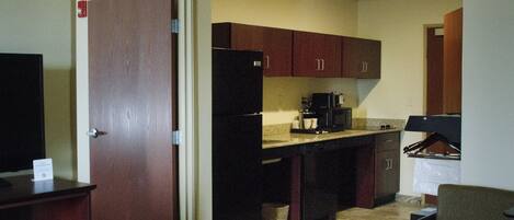 A compact kitchen with wooden cabinets, a refrigerator, a coffee maker, and a chair.