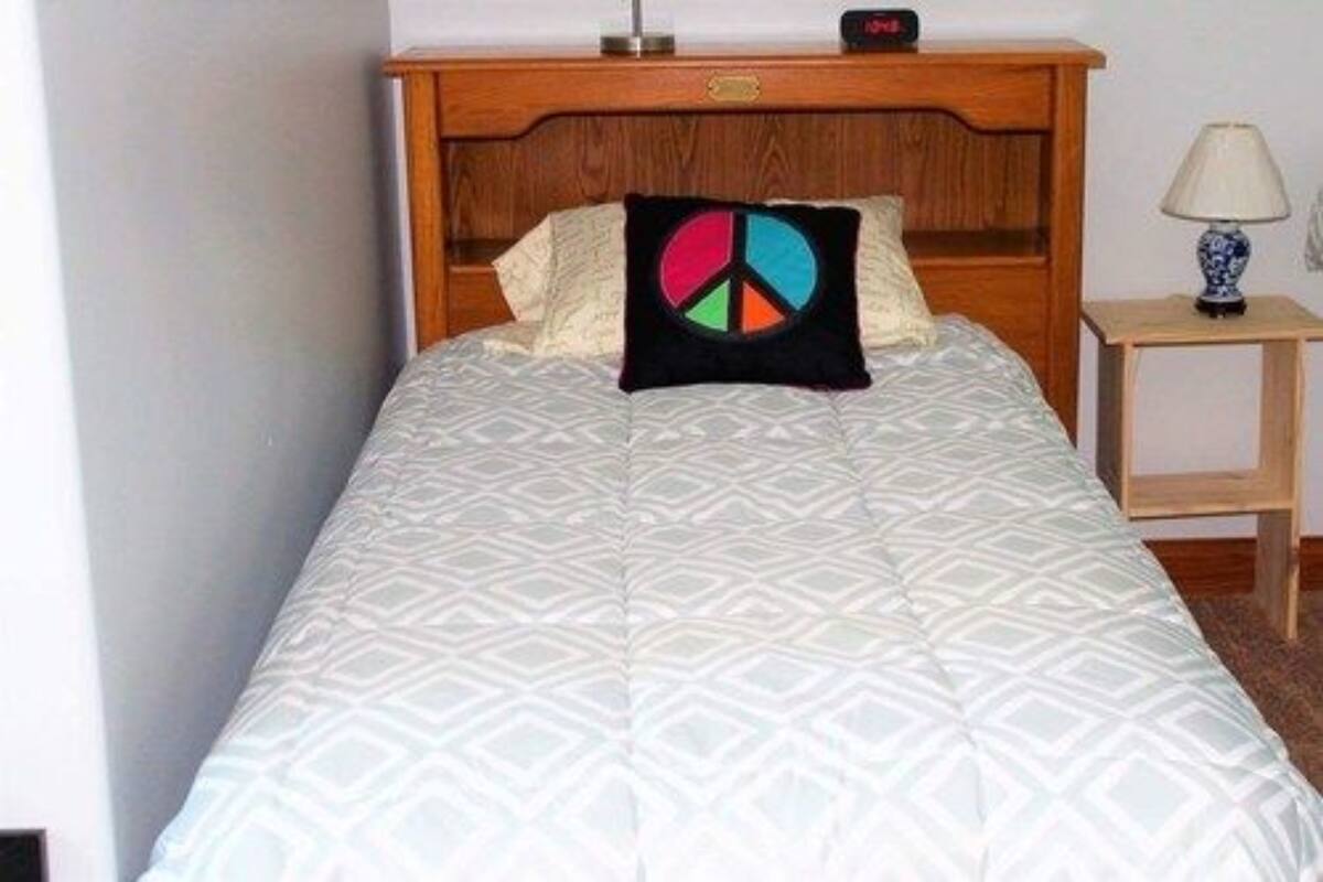 A neatly made bed with a geometric patterned bedspread and a peace sign pillow. A wooden nightstand with a lamp and a small table with a lamp is beside the bed.