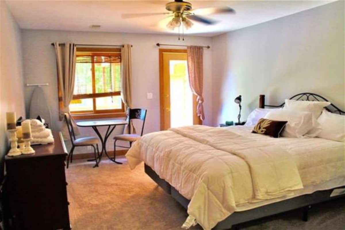 A bedroom with a bed, a desk, and a chair. There is a window with curtains, a ceiling fan, and a door.