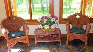Two wicker chairs with cushions, a small table with a floral arrangement, and a view of a green landscape through large windows. - Oak Creek Lodge (Bannister)