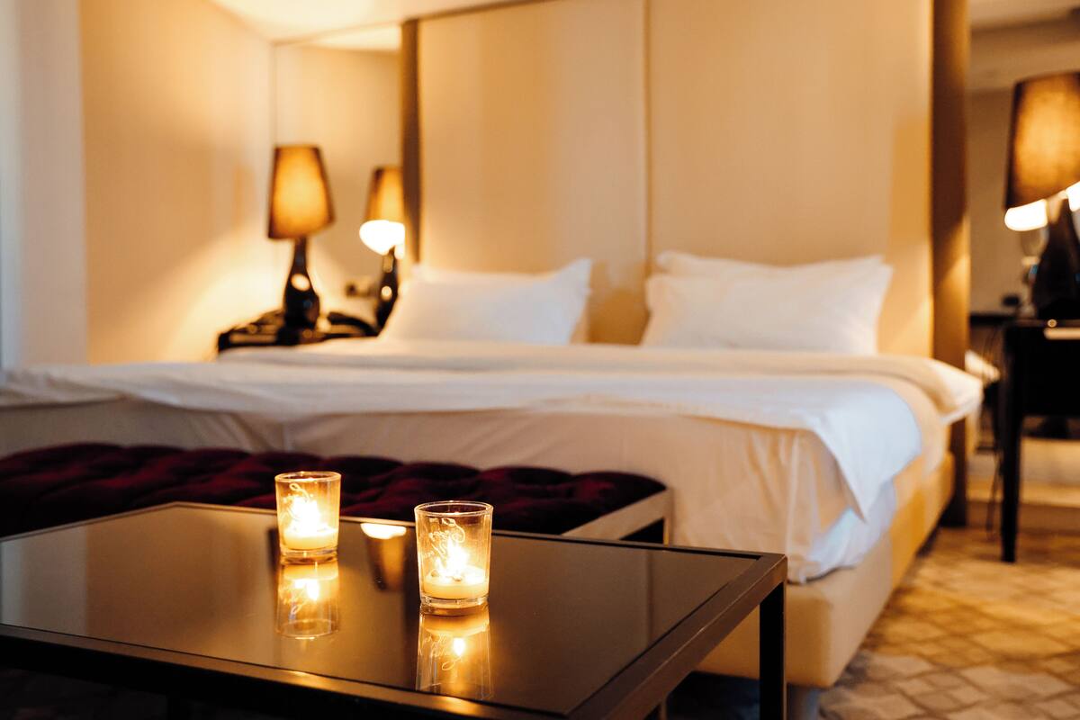 Una habitación de hotel con una cama bien hecha, dos lámparas de noche y una mesita de vidrio con velas.