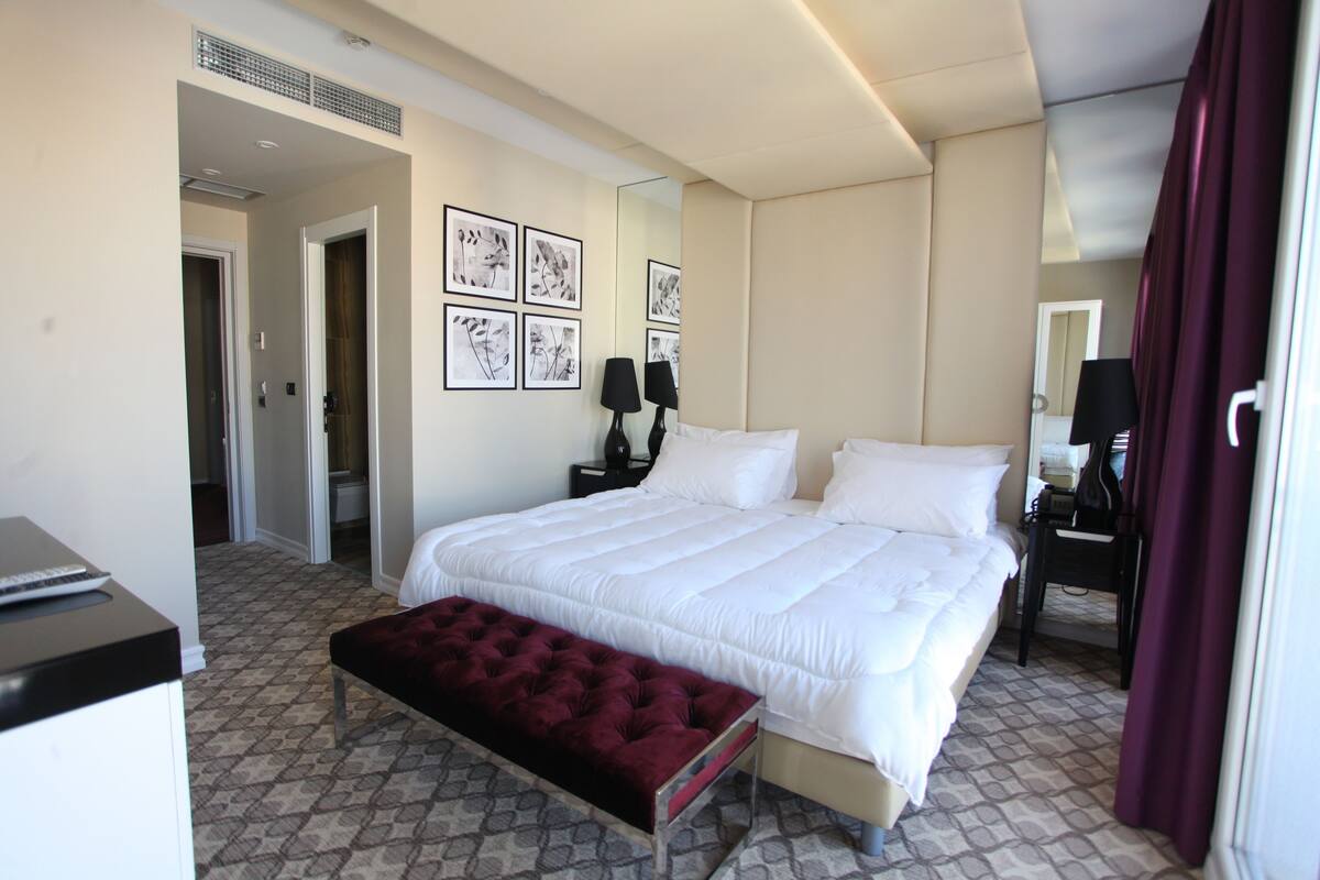 Habitación de hotel con una cama grande, una mesita de noche con lámpara, un espejo y cuadros en la pared.