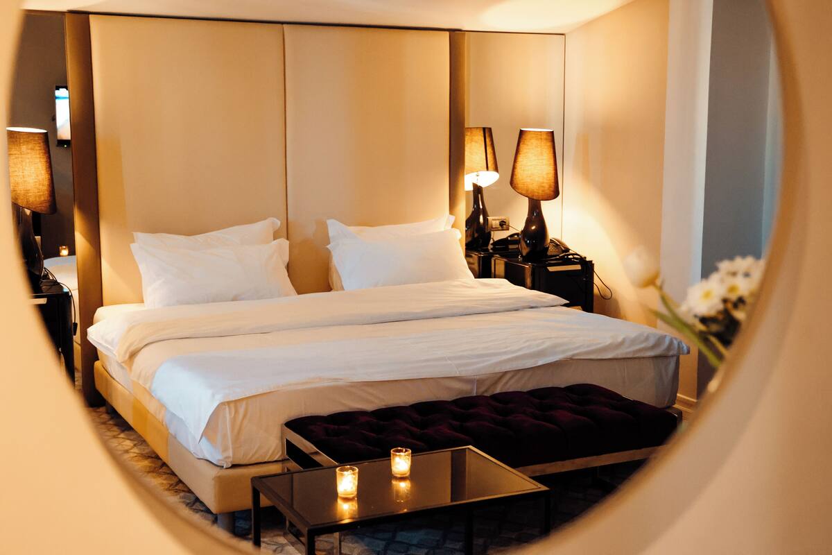 Una habitación de hotel con una cama grande, lámparas de noche, una mesita pequeña y un otomano oscuro.