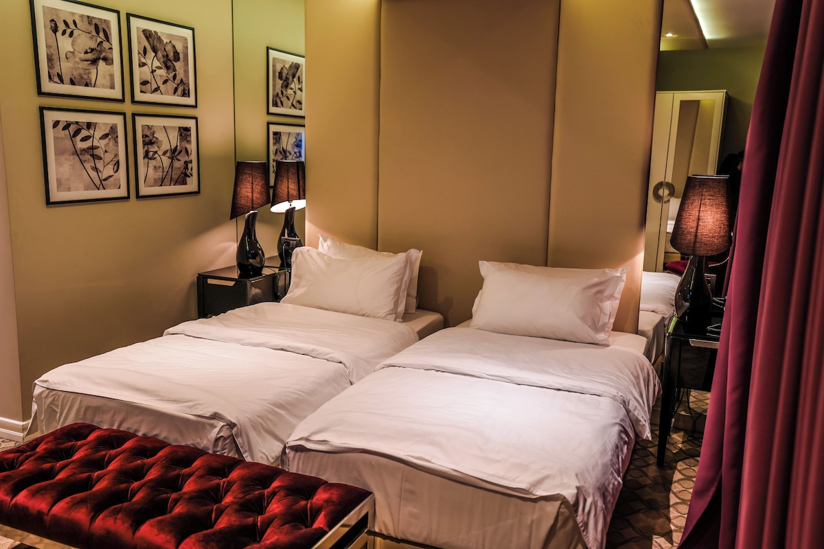 Une chambre d’hôtel avec deux lits, un banc en velours rouge et des œuvres d’art accrochées au mur.