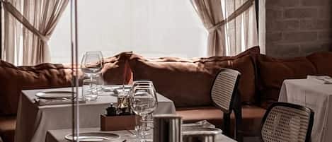 Un coin repas avec une table dressée pour deux personnes, un banc rembourré et une vue par la fenêtre, qui est agrémentée de rideaux.
