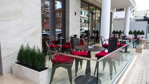 An outdoor seating area with tables and chairs, a planter with greenery, and a glass railing. - Senator Hotel (Tirana)