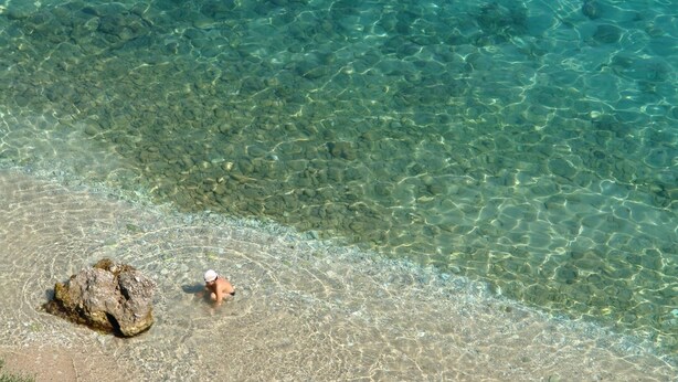 Una vista despejada de una playa con una persona en el agua y una roca grande en la orilla.