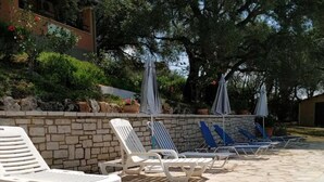 Un patio de piedra con sillones de descanso y sombrillas, rodeado de follaje y un muro de piedra.