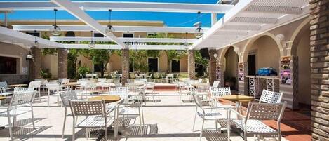An outdoor patio area with white metal chairs and tables, a pergola structure, and a brick wall.