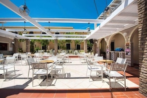 Un patio al aire libre con sillas y mesas de metal blanco, una estructura de pérgola y un muro de ladrillo.