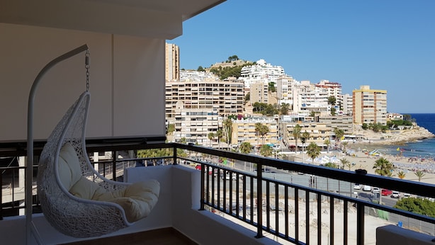 Uma varanda com uma cadeira suspensa, vista para uma praia e um horizonte urbano ao fundo.