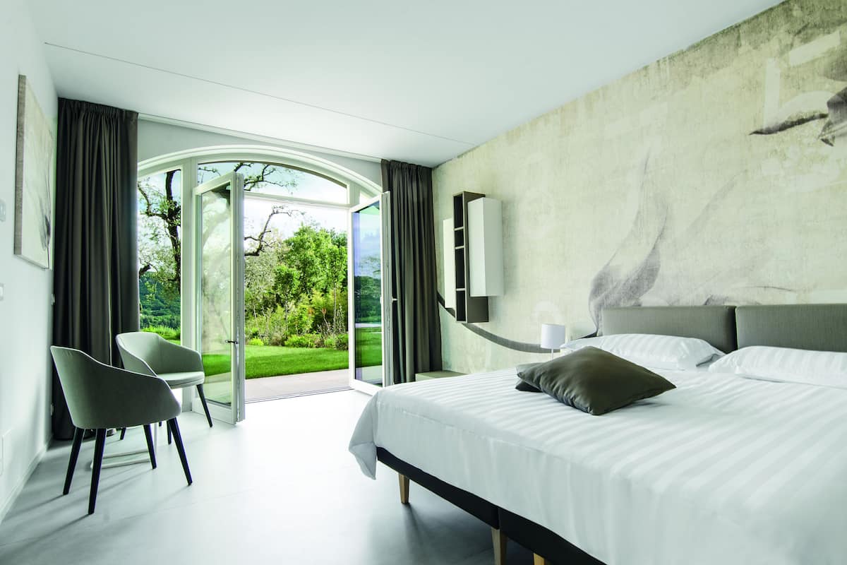 A modern bedroom with a bed, a chair, and a view of a garden through a glass door.
