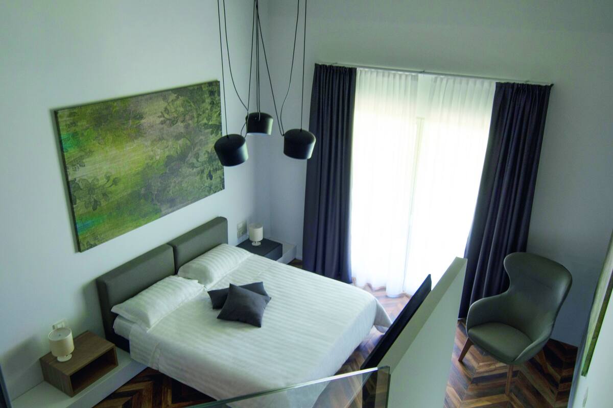 A hotel room with a bed, a chair, a nightstand, a lamp, and a painting on the wall.