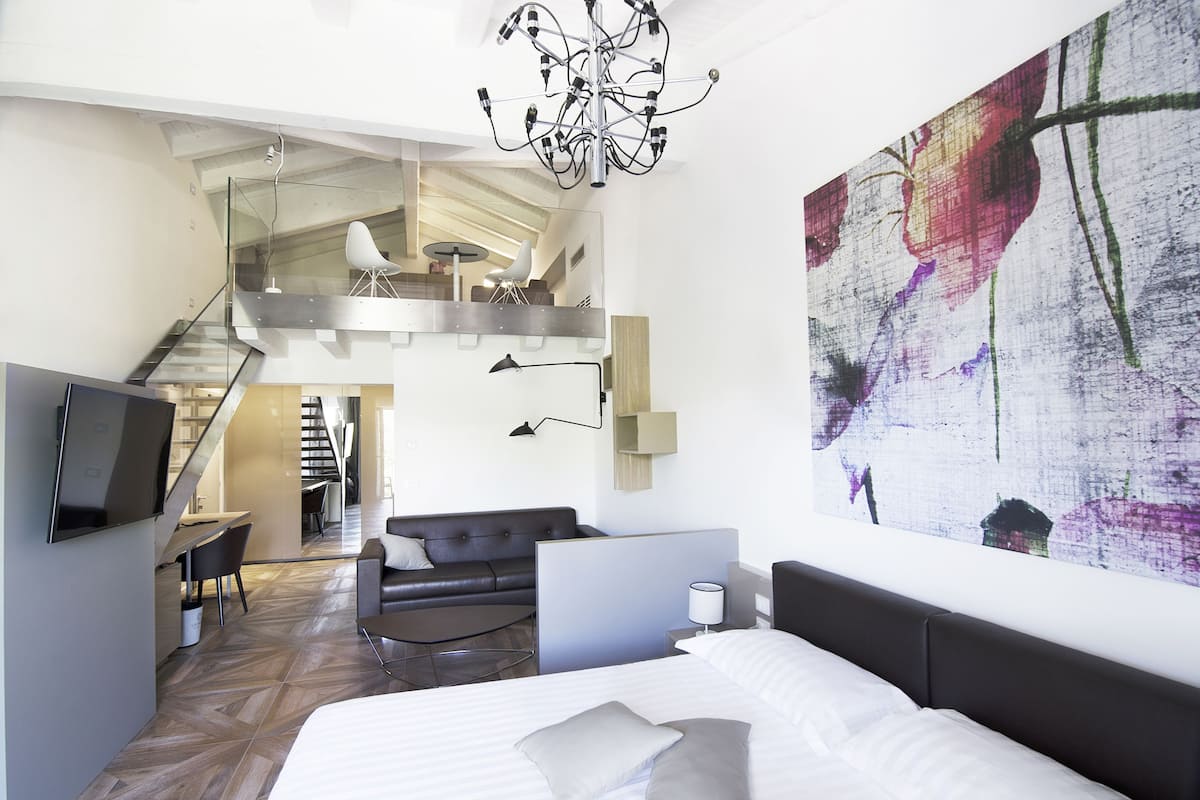 A modern hotel room with a large bed, a desk, a laptop, a sofa, and a staircase leading to an upper level.