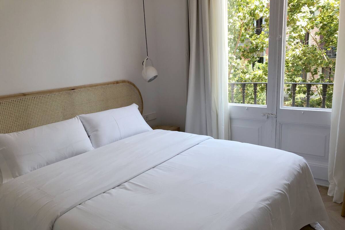 Un lit bien fait, avec du linge de lit blanc, dans une chambre dotée d’une tête de lit en bois, d’une lampe suspendue et offrant une vue sur la verdure par la fenêtre.