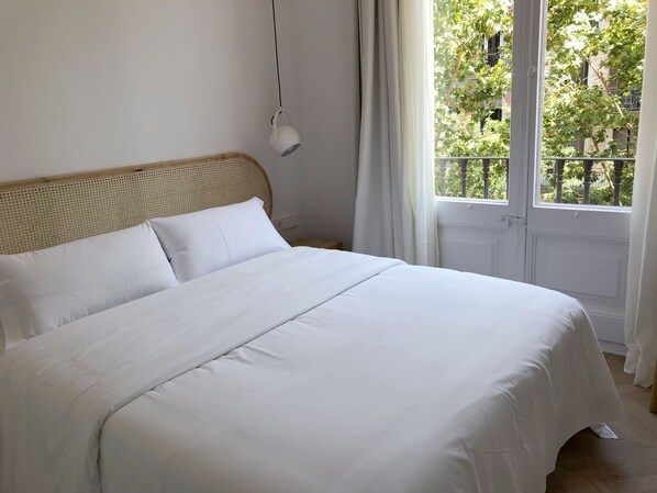 Una cama bien hecha con sábanas blancas en una habitación con cabecera de madera, una lámpara colgante y vistas a zonas verdes por la ventana.