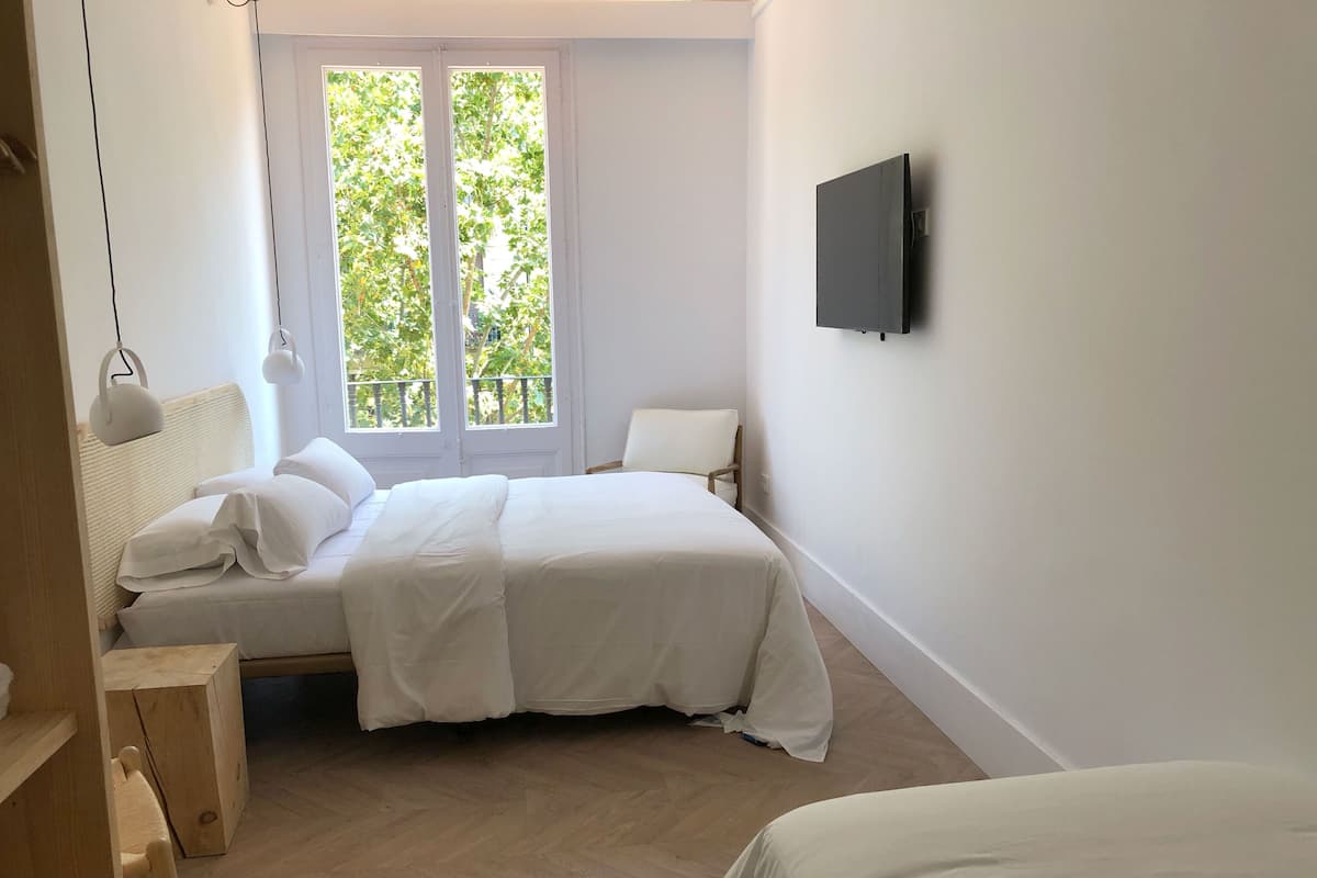 Une chambre à coucher avec un lit, une chaise, un téléviseur fixé au mur et une fenêtre donnant sur des arbres.