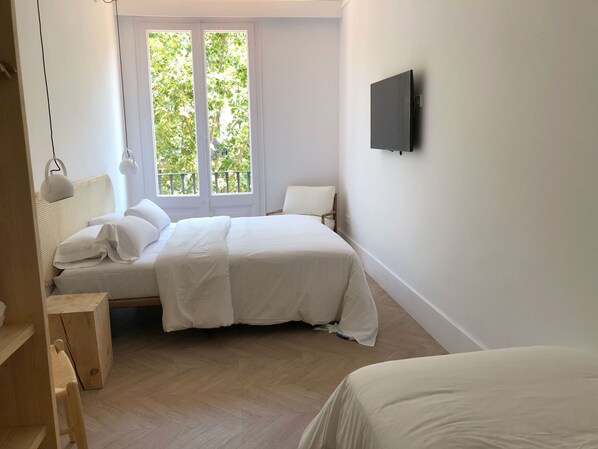 Un dormitorio con cama, una silla, televisor en la pared y ventana con vistas a árboles.