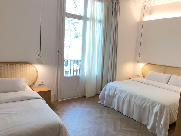A hotel room with two beds, a wooden headboard, a small bedside table, a nightstand, a window with curtains, and a wall-mounted light fixture. - The Mohouse Boutique Room (Barcelona)