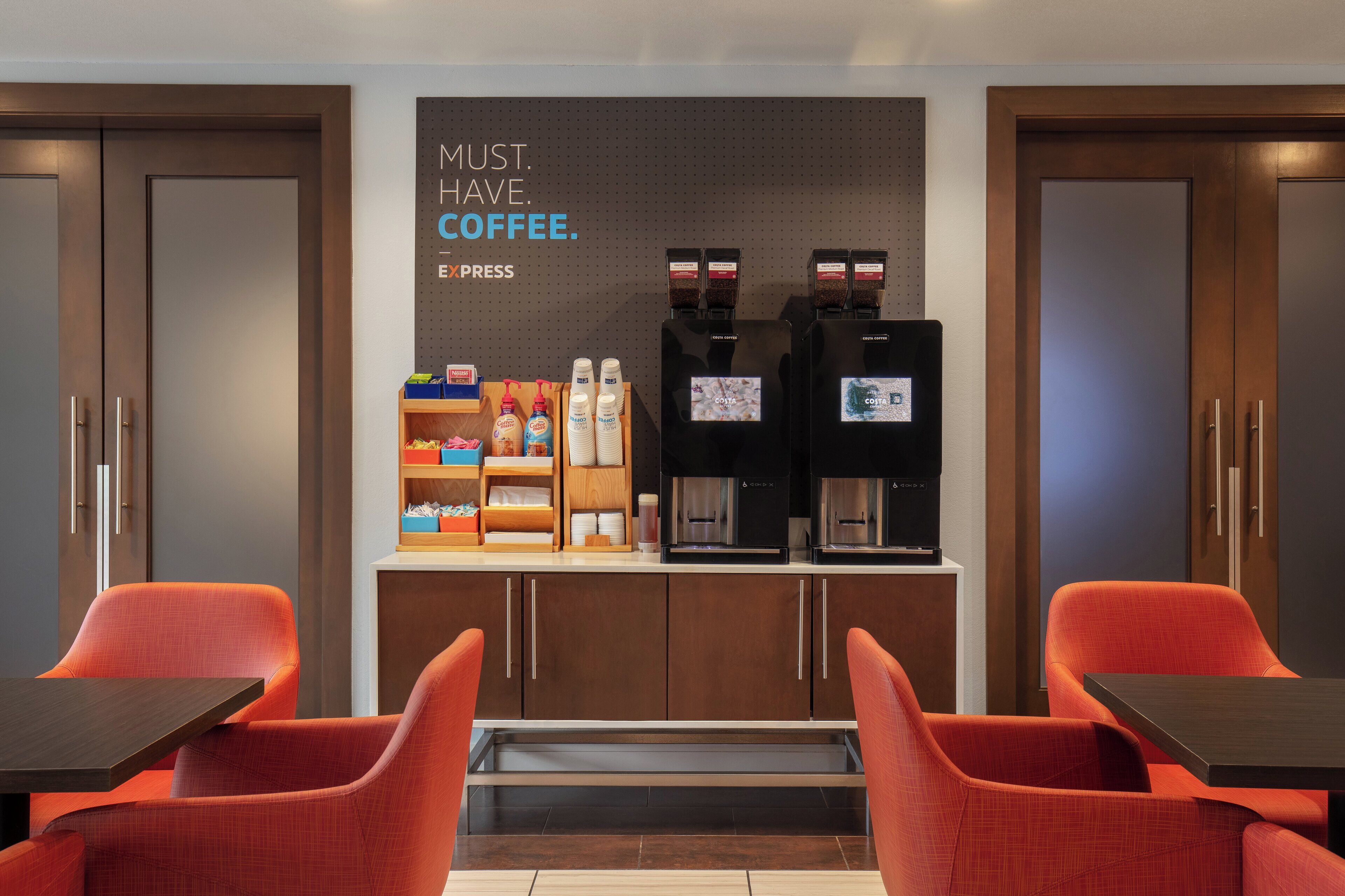A coffee station with two espresso machines, a coffee display, and a seating area with two orange chairs and a table.