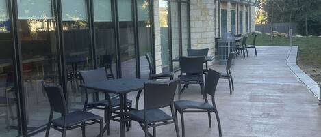Une terrasse extérieure avec des tables et des chaises, un bâtiment en pierre doté de grandes fenêtres et un chemin piétonnier en béton.