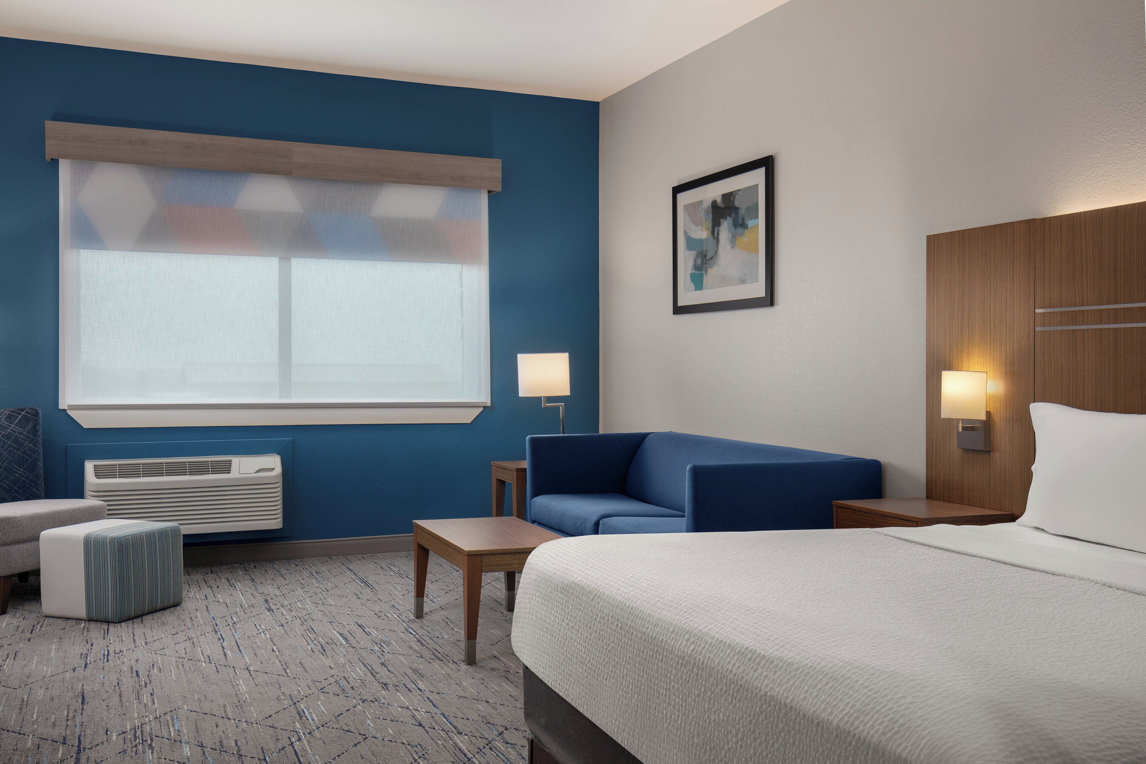 A hotel room with a bed, a blue sofa, a small table, a lamp, and a window with blinds.