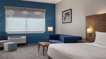 Une chambre d’hôtel avec un lit, un canapé bleu, une petite table, une lampe et une fenêtre avec des stores.