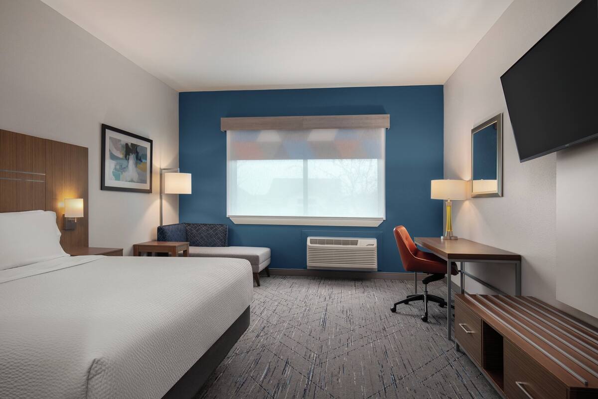 A hotel room with a large bed, a desk, a chair, a small table, a lamp, a TV, and a window with blinds.