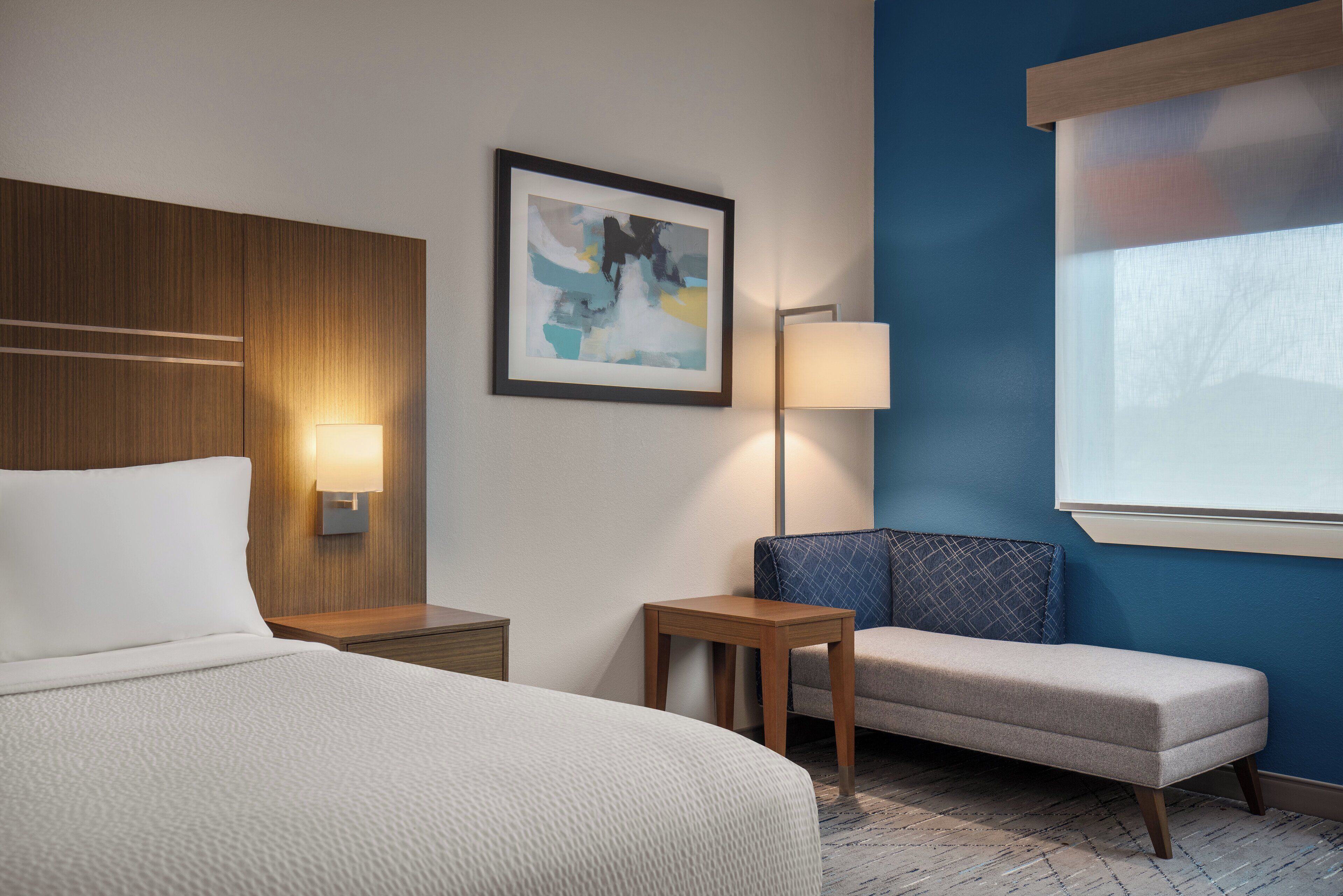 Habitación de hotel con cama, una mesita, una silla, una lámpara y un cuadro en la pared.
