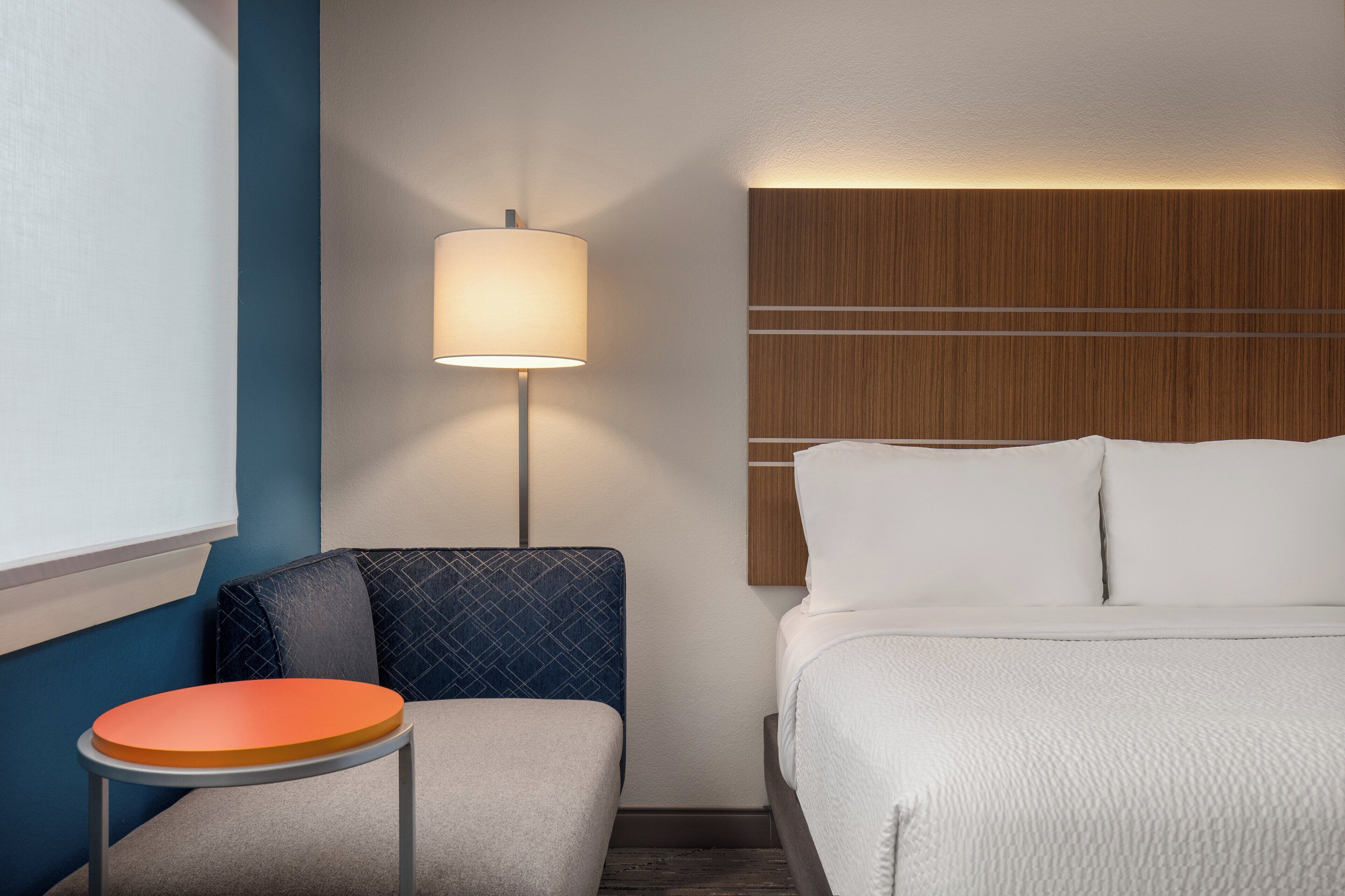 A hotel room with a bed, a nightstand with a lamp, a small table, and a blue wall.