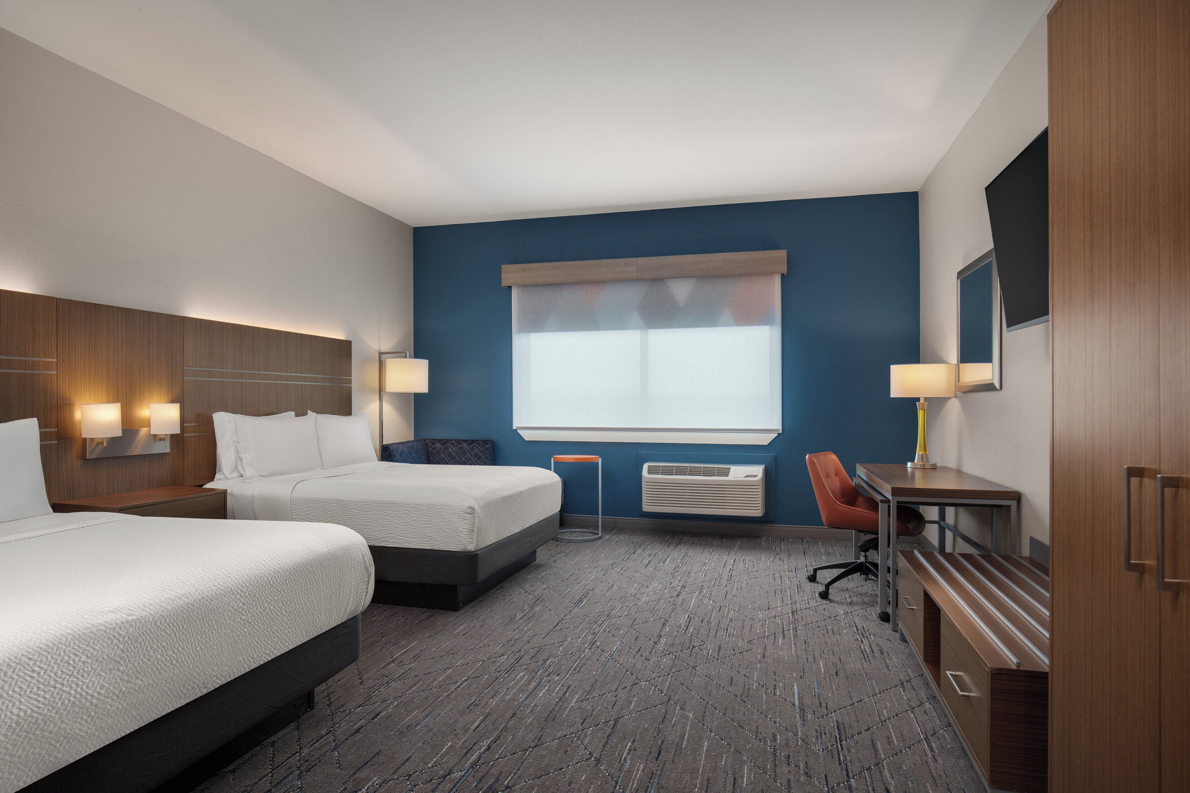 A hotel room with two beds, a desk, a chair, a lamp, a mirror, and a window with blinds.