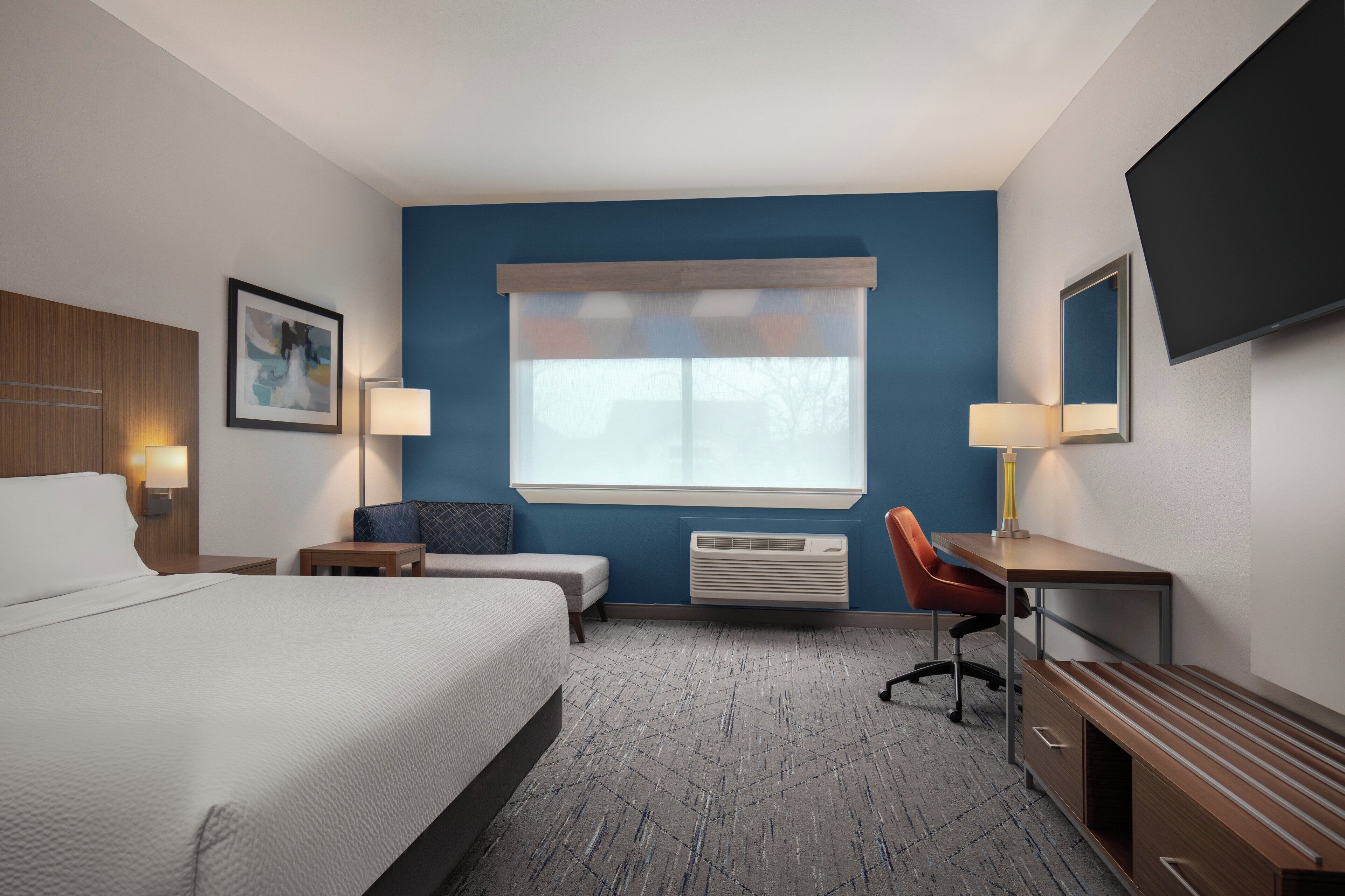 A hotel room with a large bed, a desk, a chair, a small table, a lamp, a TV, and a window with blinds.