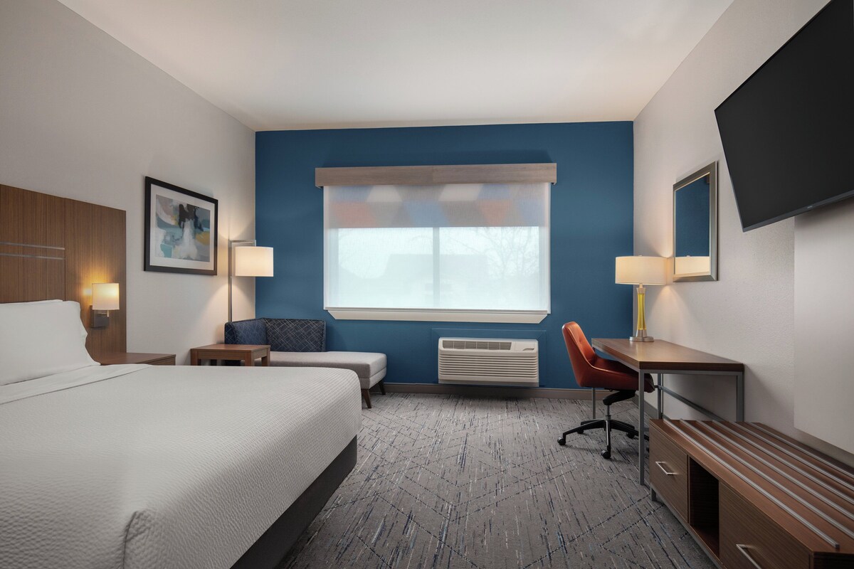 A hotel room with a large bed, a desk, a chair, a small table, a lamp, a TV, and a window with blinds.