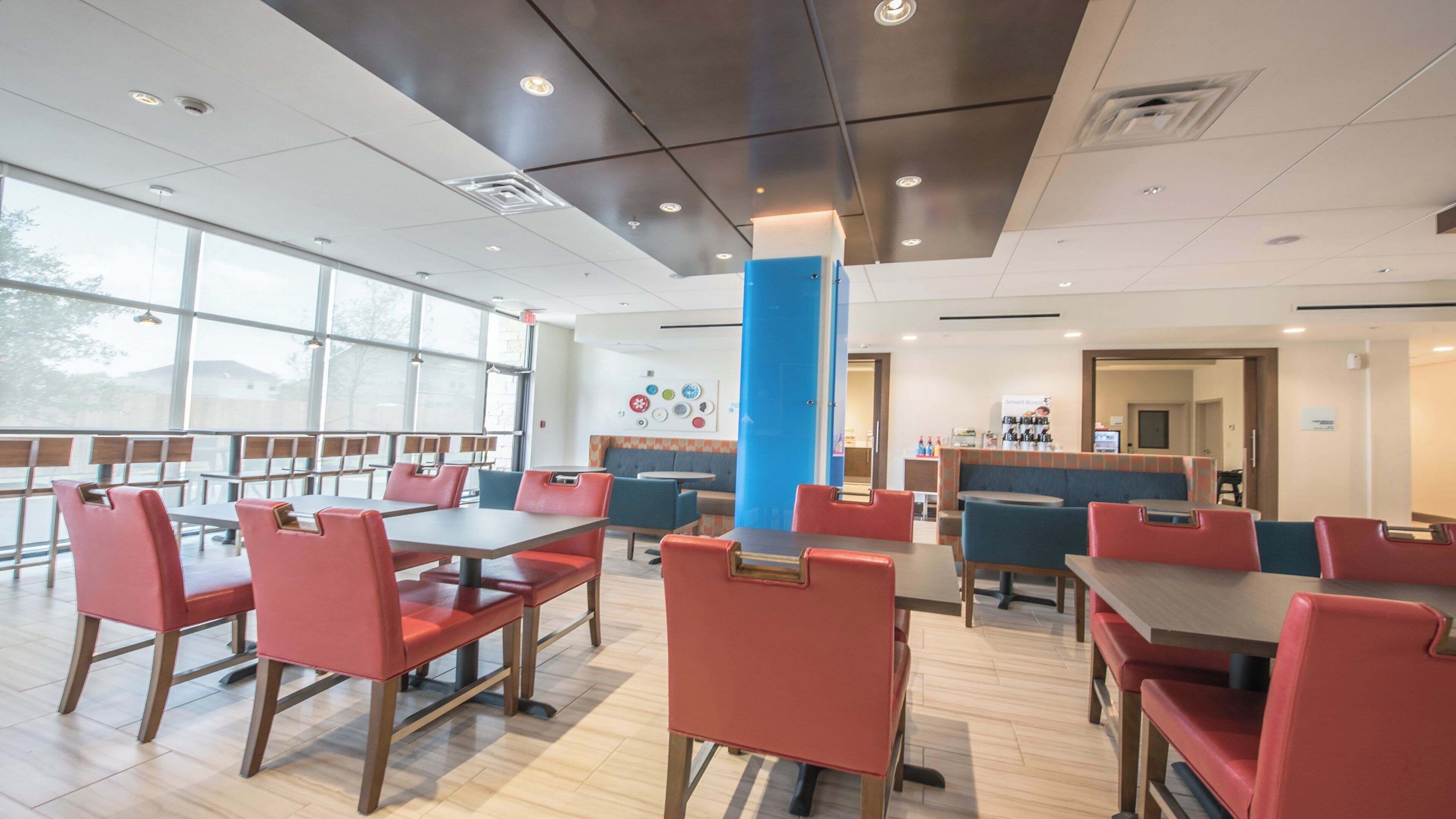 A modern dining area with red chairs and tables, large windows, and a blue pillar.