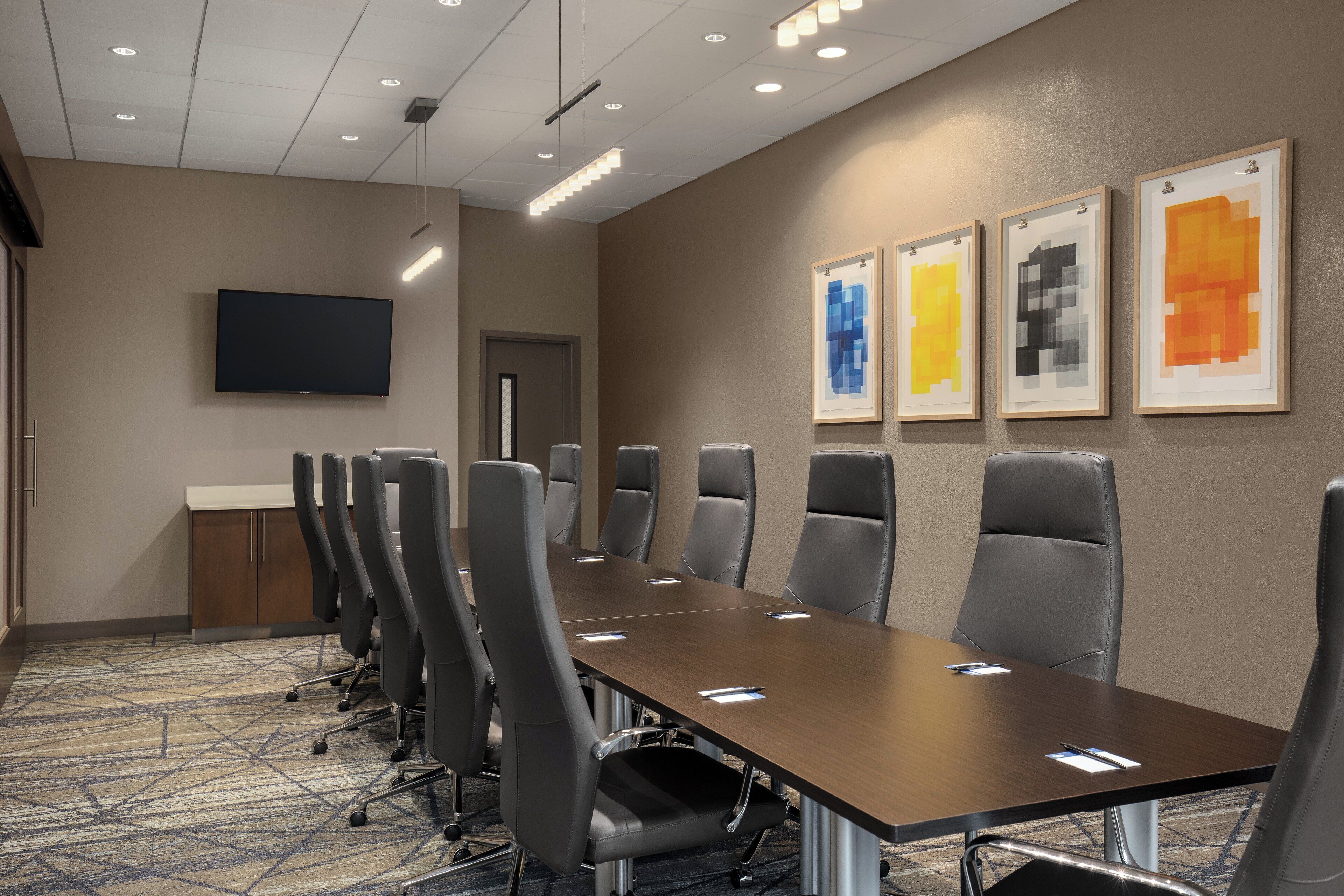 A conference room with a long table and multiple chairs, a flat-screen TV mounted on the wall, and three framed artworks on the wall.