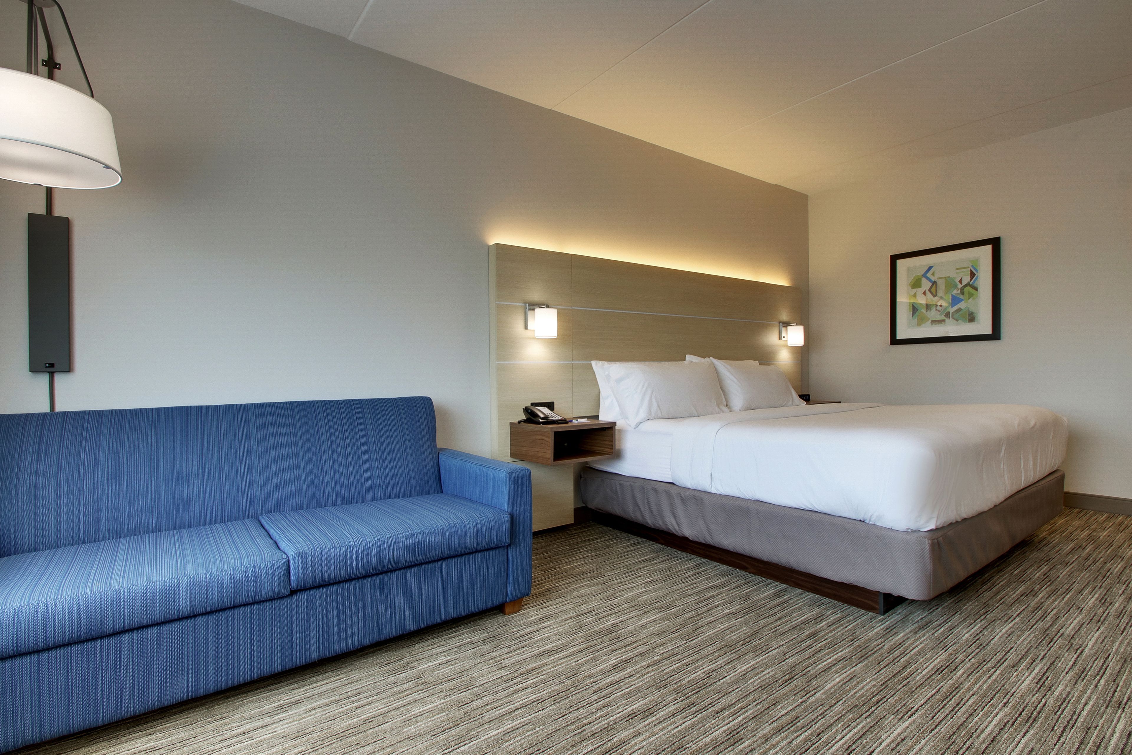 A hotel room with a blue sofa, a bed with white linens, a nightstand with a lamp, and a framed picture on the wall.