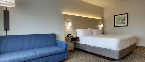 A hotel room with a blue sofa, a bed with white linens, a nightstand with a lamp, and a framed picture on the wall.