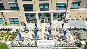 An outdoor patio with multiple tables and chairs, a glass entrance, and a brick building facade.
