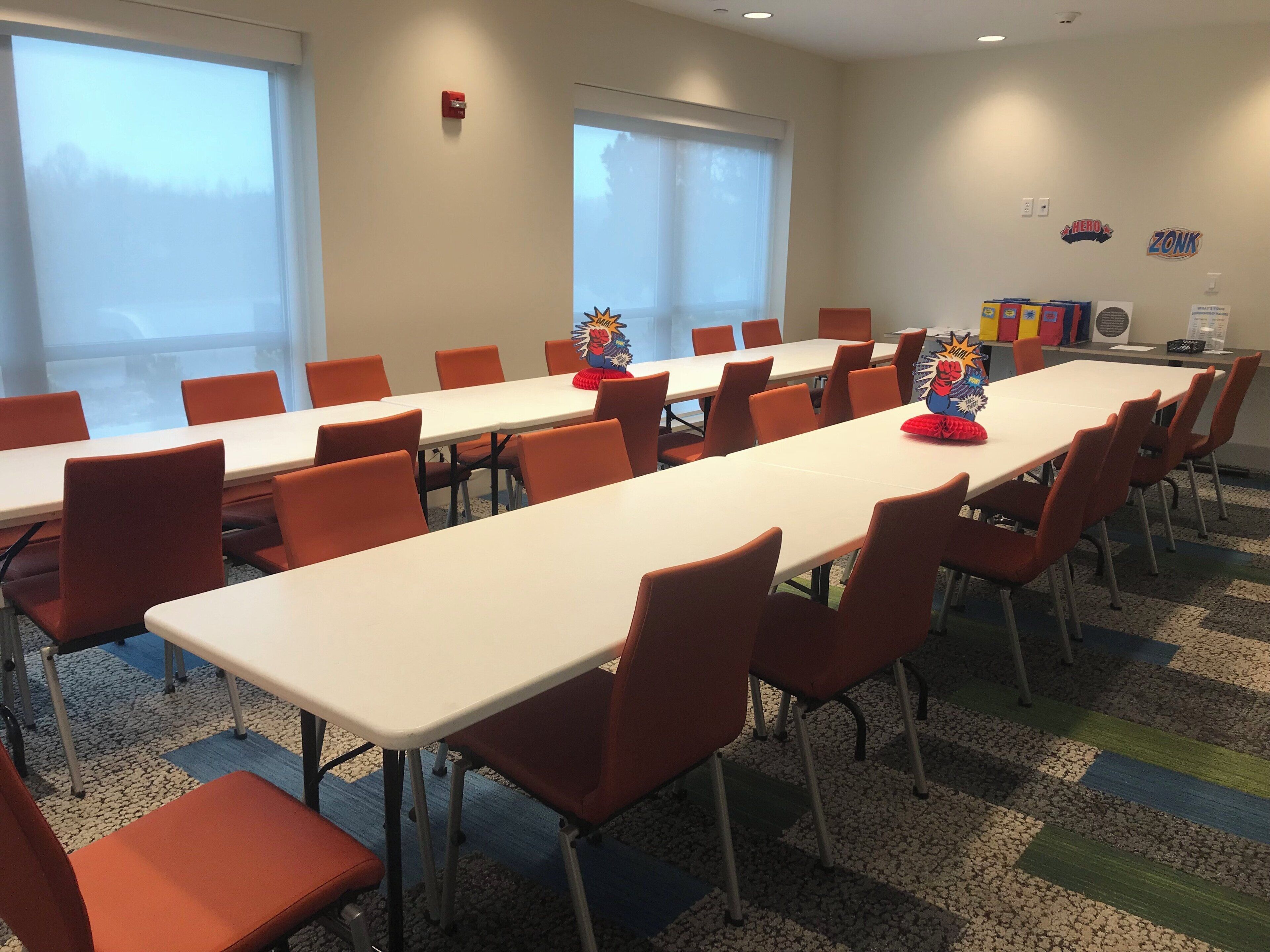 A conference room with long tables and orange chairs, large windows, and a fire alarm on the wall.