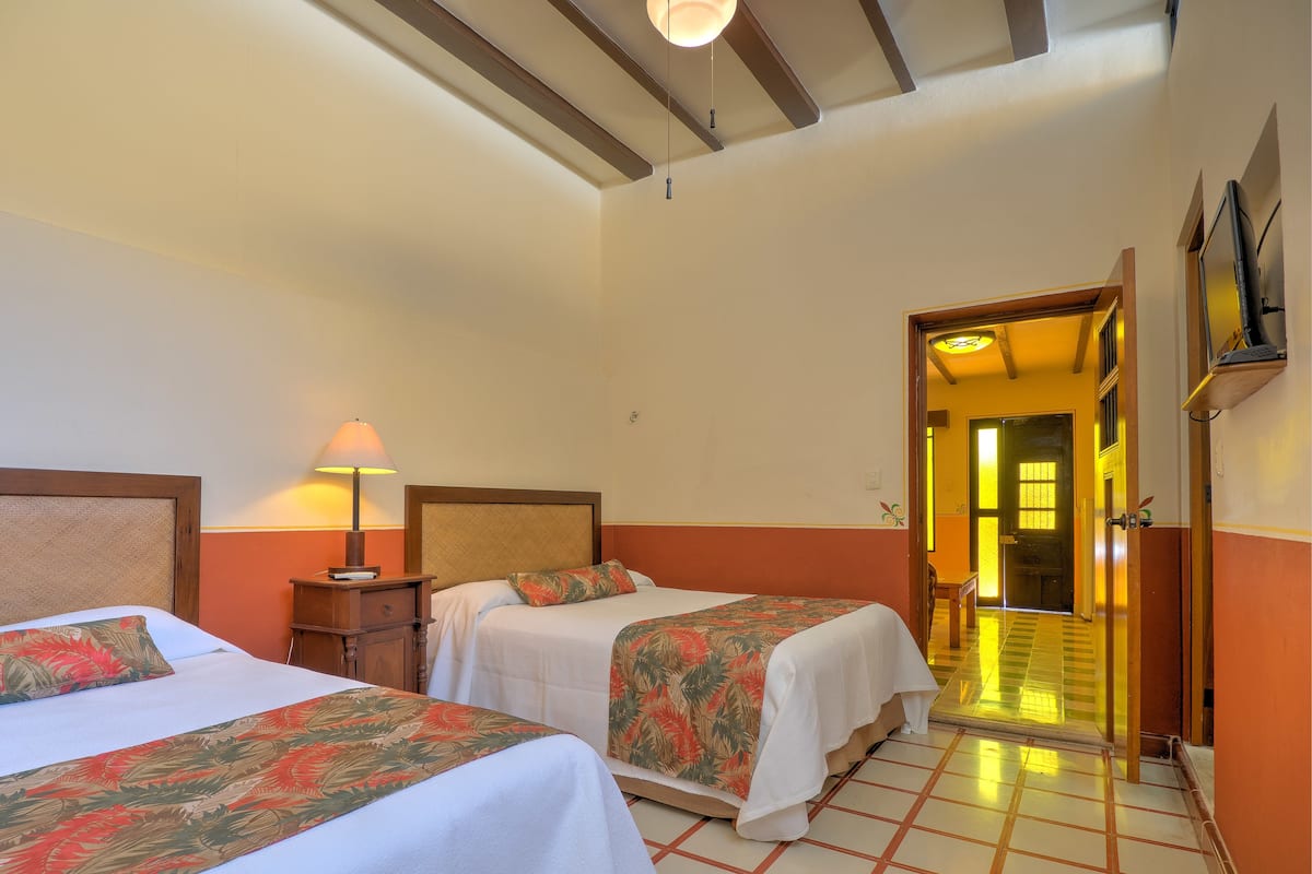 Habitació d'hotel amb dos llits, una tauleta de nit, una làmpada i una porta que dona a una altra habitació.