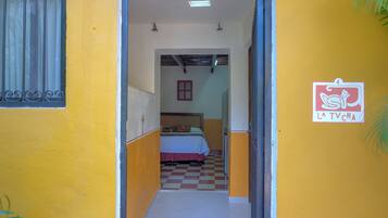Um corredor estreito conduz a um quarto com cama e piso xadrez. Uma placa com um personagem de desenho animado e texto é visível na parede.