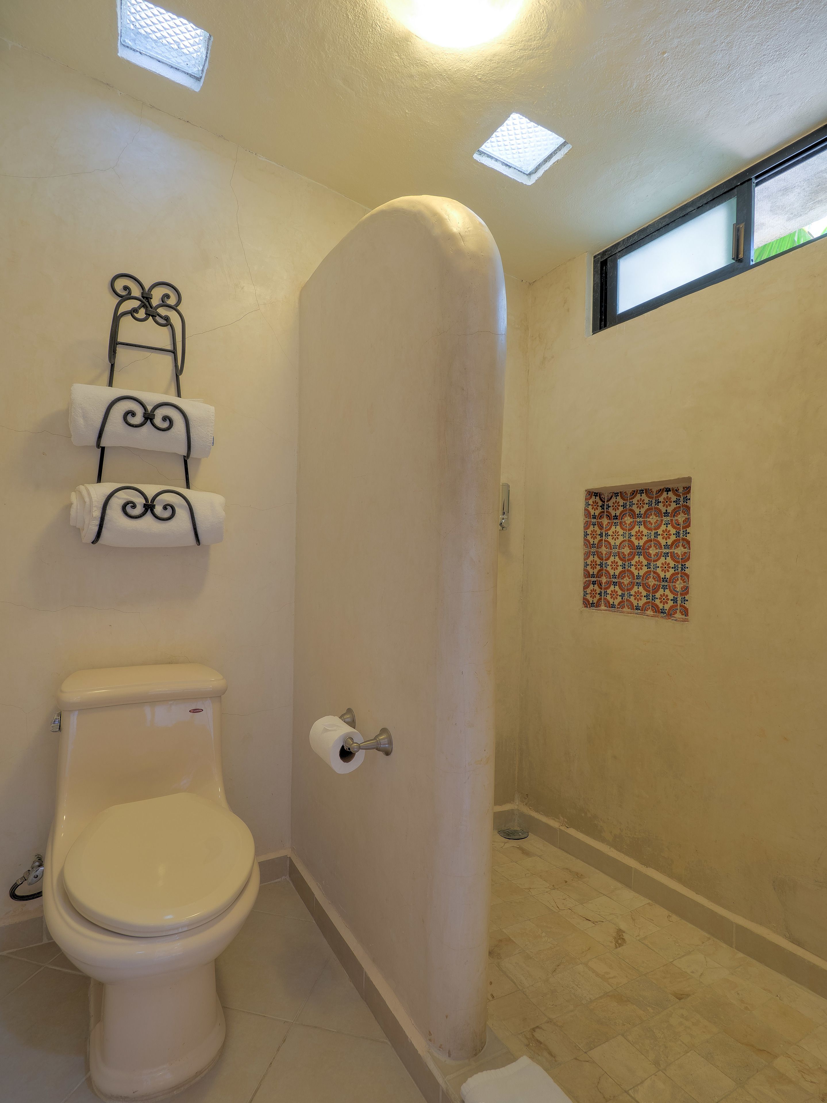 A bathroom with a toilet, a towel rack with three towels, a shower door, and a small window.