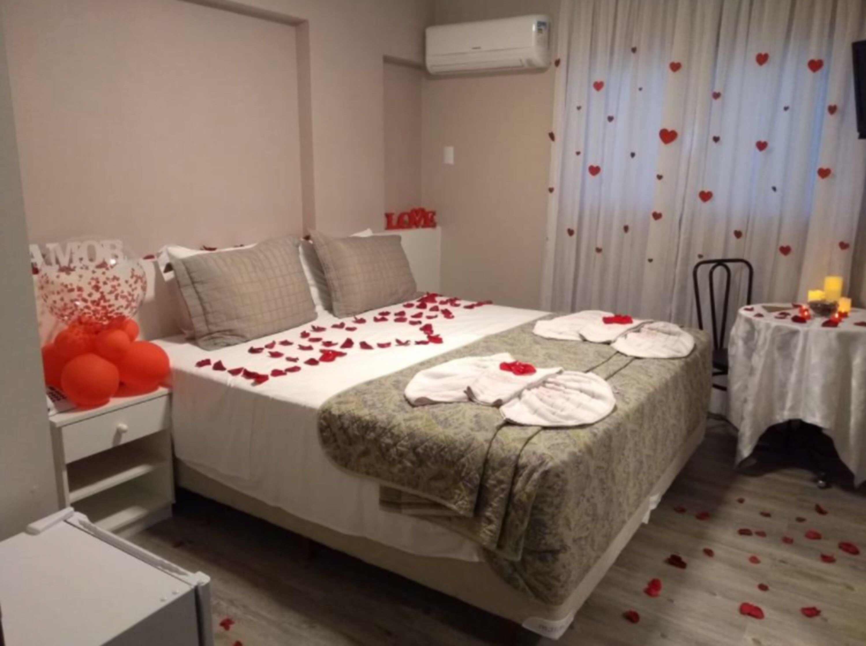 Habitación de hotel con cama, mesita de noche, pequeña mesa con velas y cortina con diseño de corazones.