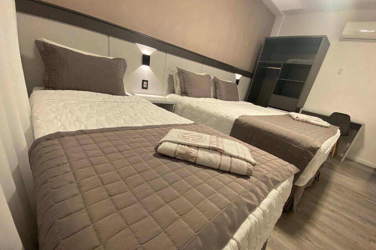 A hotel room with two beds, each with a quilted bedspread and pillows. There is a wardrobe and a small table with a lamp.