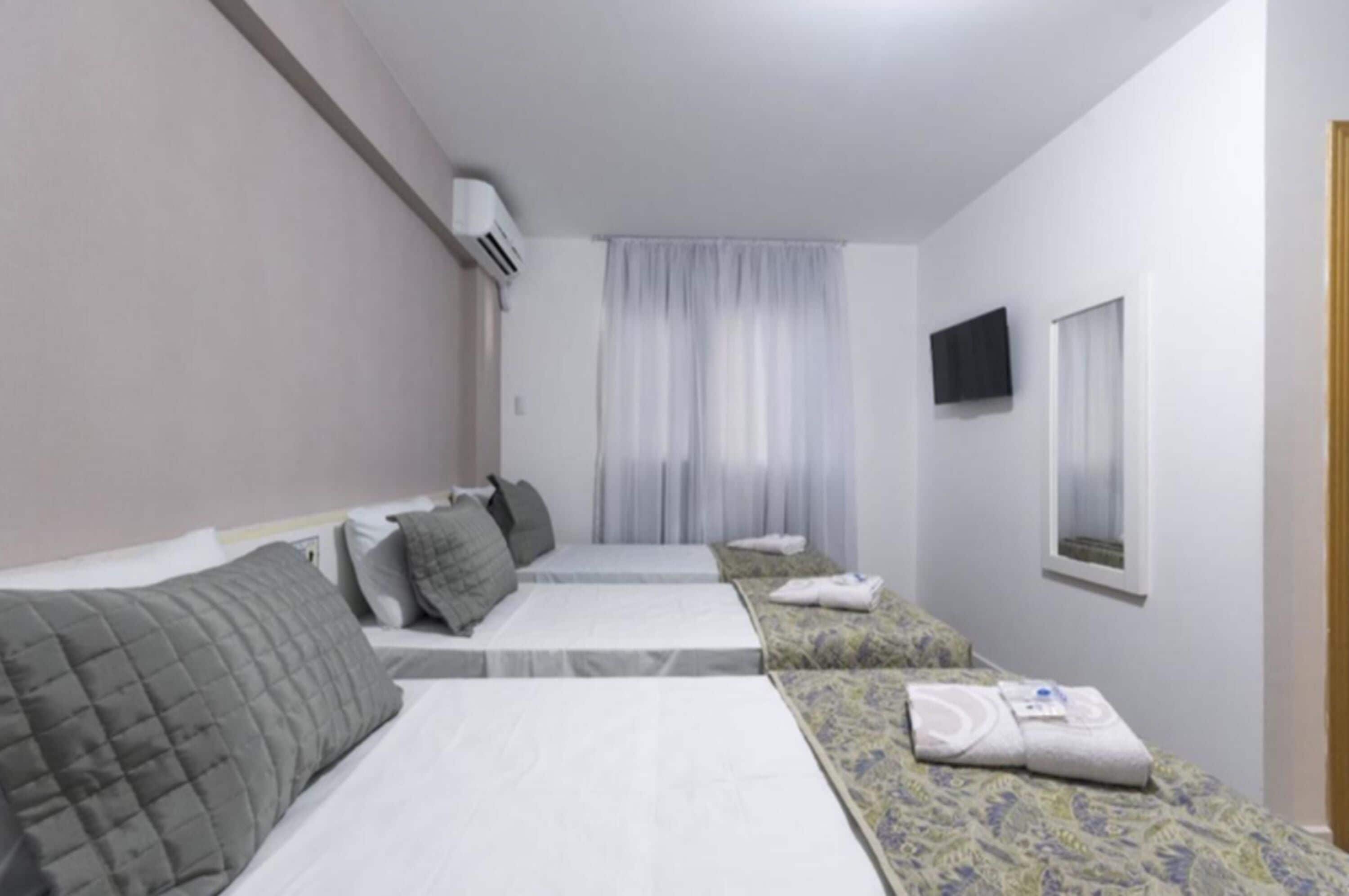 A hotel room with two beds, a flat-screen TV, a window with curtains, and an air conditioning unit.