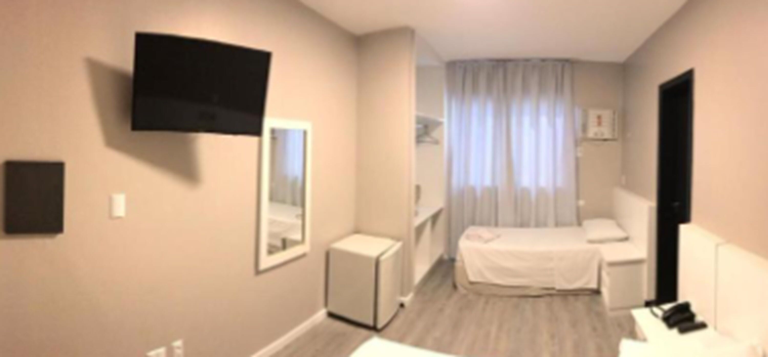 A hotel room with a bed, a TV mounted on the wall, a small bedside table, and a window with curtains.