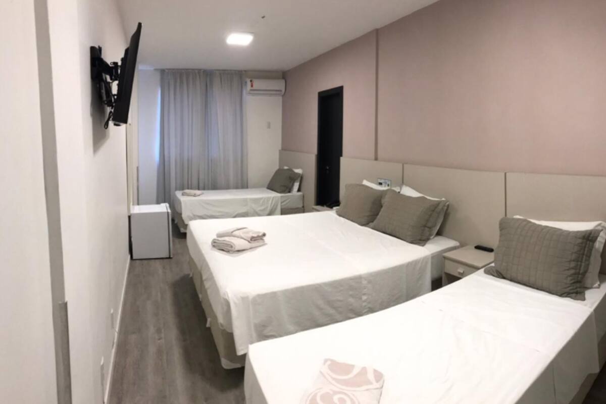 A hotel room with three single beds, a small bedside table with a lamp, a flat-screen TV mounted on the wall, and a window with curtains.